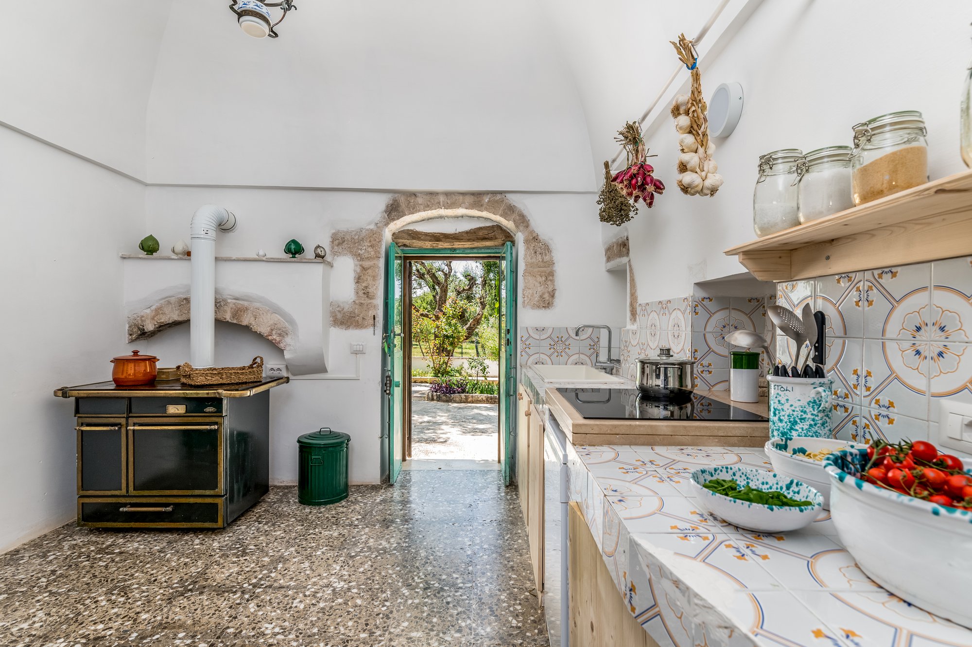 Stone kitchen with a wood-burning stove, Apulian ceramics and a green door onto the garden of the masseria